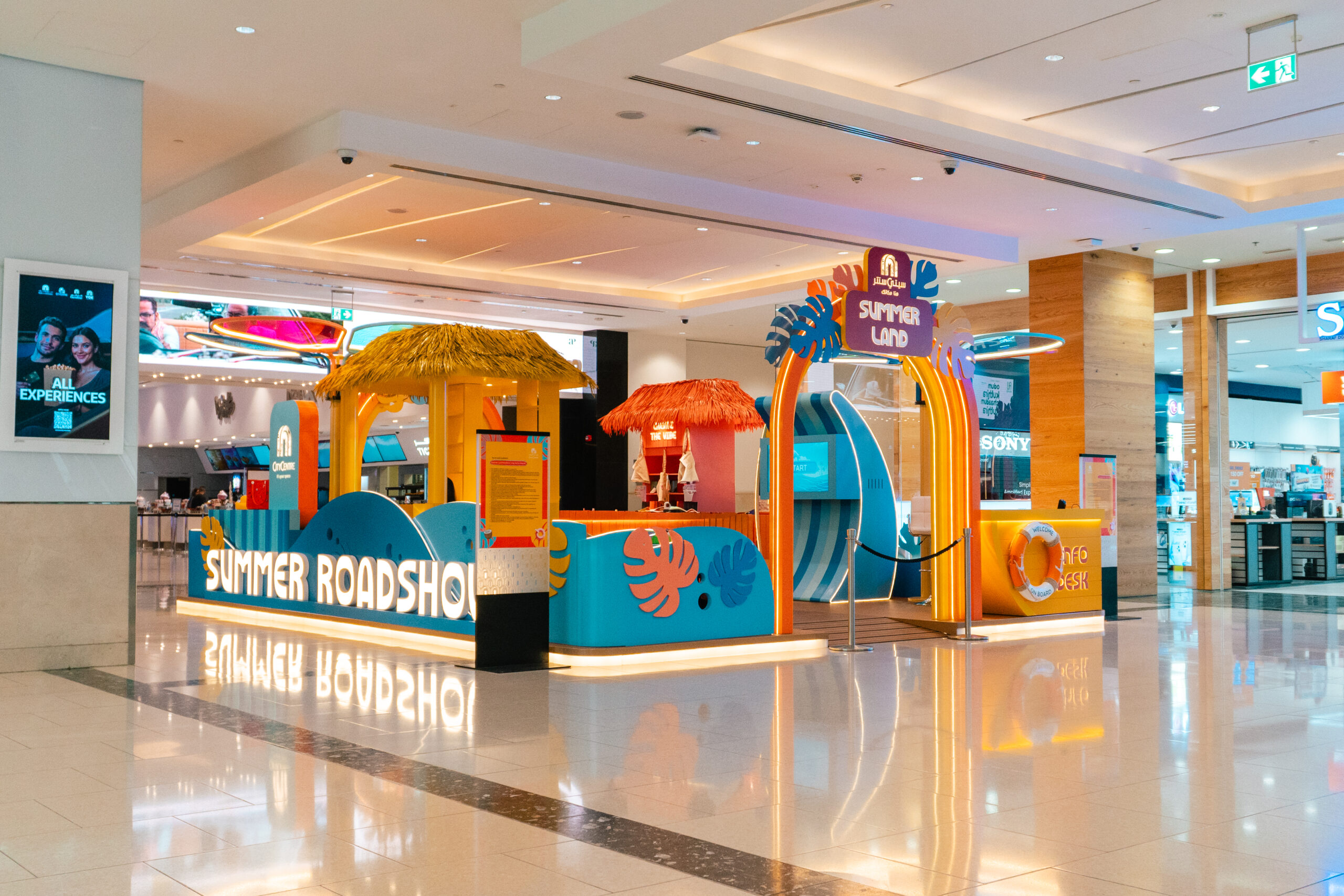 The Summer Roadshow activation stand in the mall concourse, in orange and blue with a wave-shaped canopy