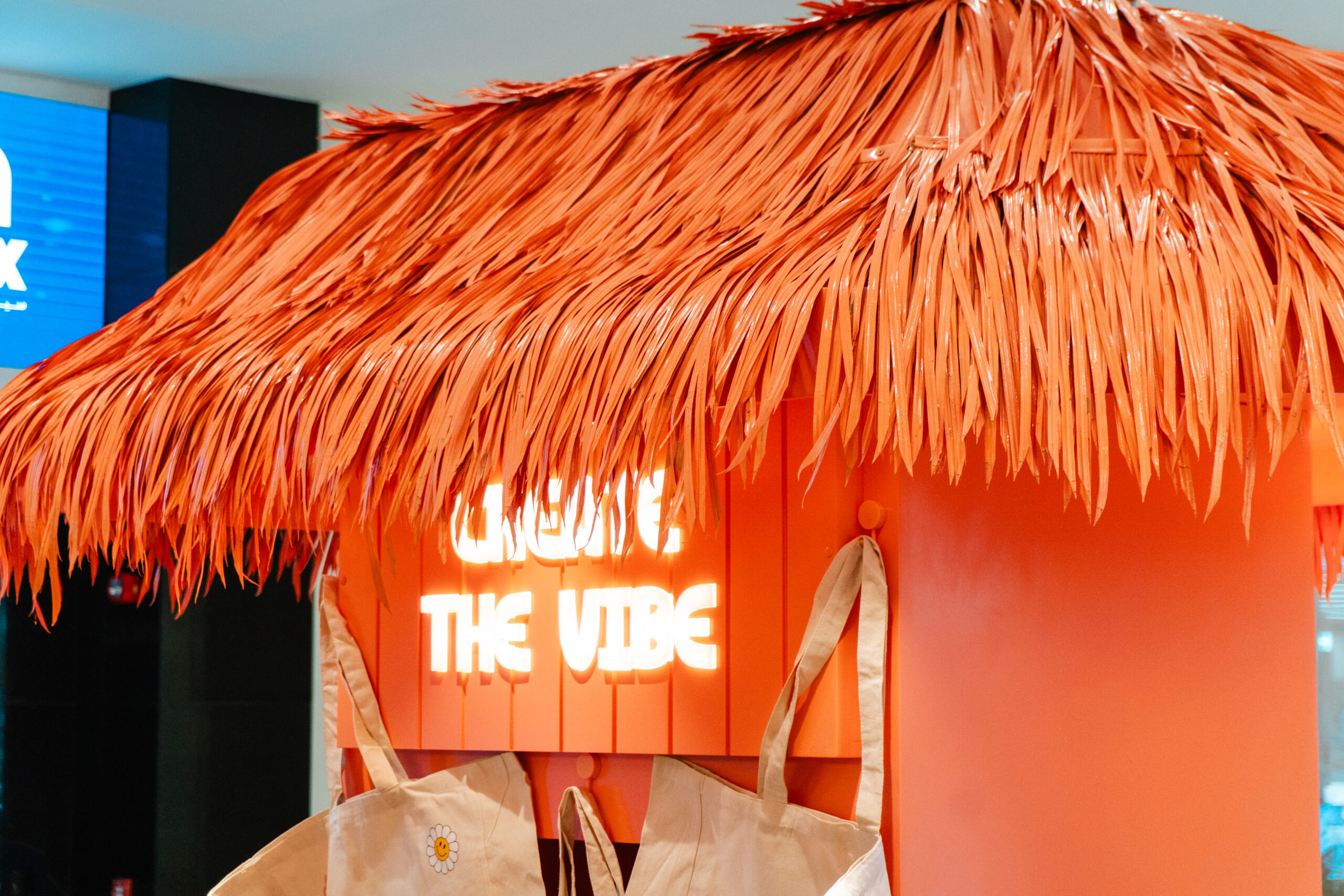 A thatched tiki booth with a neon "Create the Vibe" sign above tote bags ready for customising