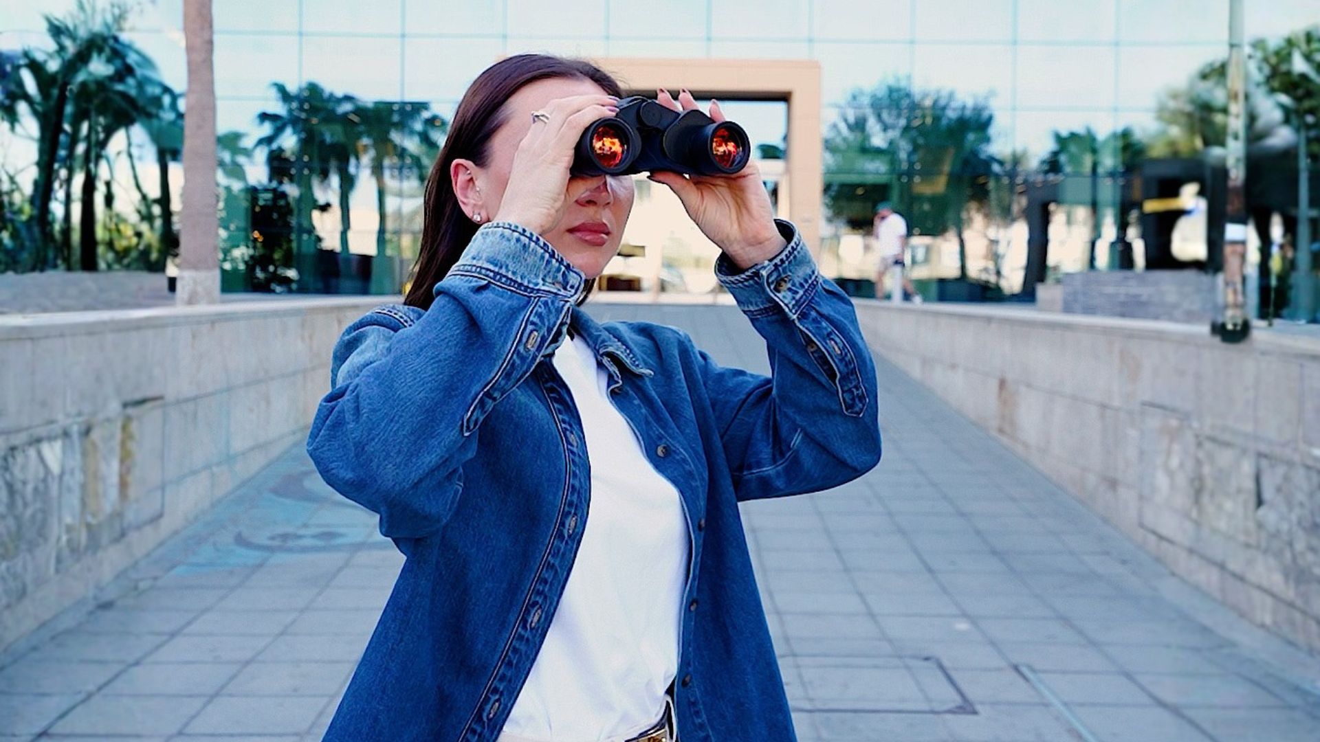 A woman in a denim jacket raising binoculars on the mall's palm-lined entrance walkway, mirrored glass facade behind her