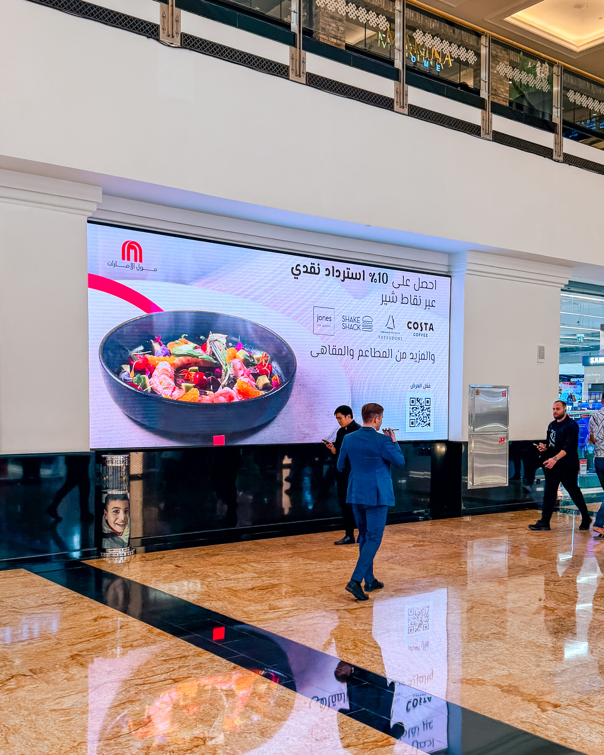 A large Arabic-language SHARE Points cashback wall screen with shoppers passing on the polished floor.