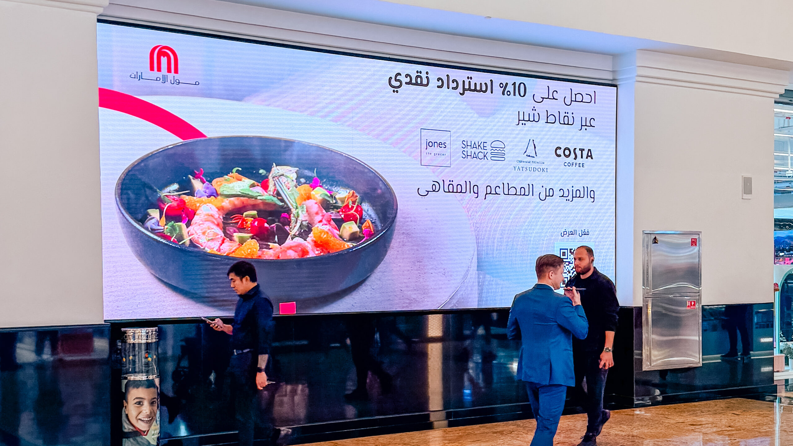 Shoppers pausing in front of the Arabic SHARE Points cashback screen at Mall of the Emirates.
