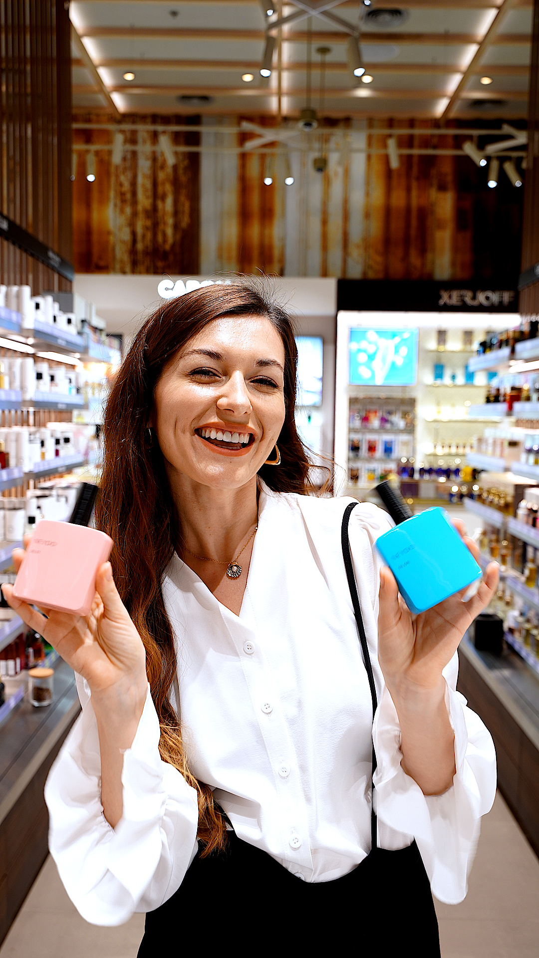 A smiling presenter in a white blouse holding up a pink and a blue product box in the fragrance hall