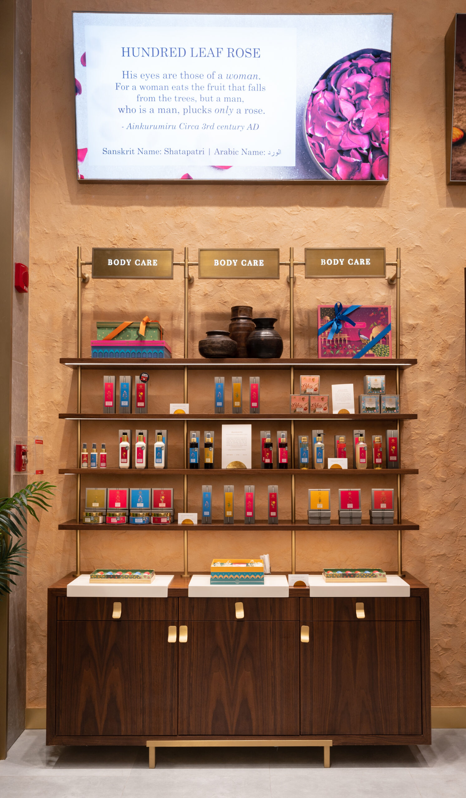 Forest Essentials body care display: brass shelving of boxed gift sets beneath a Hundred Leaf Rose ingredient screen