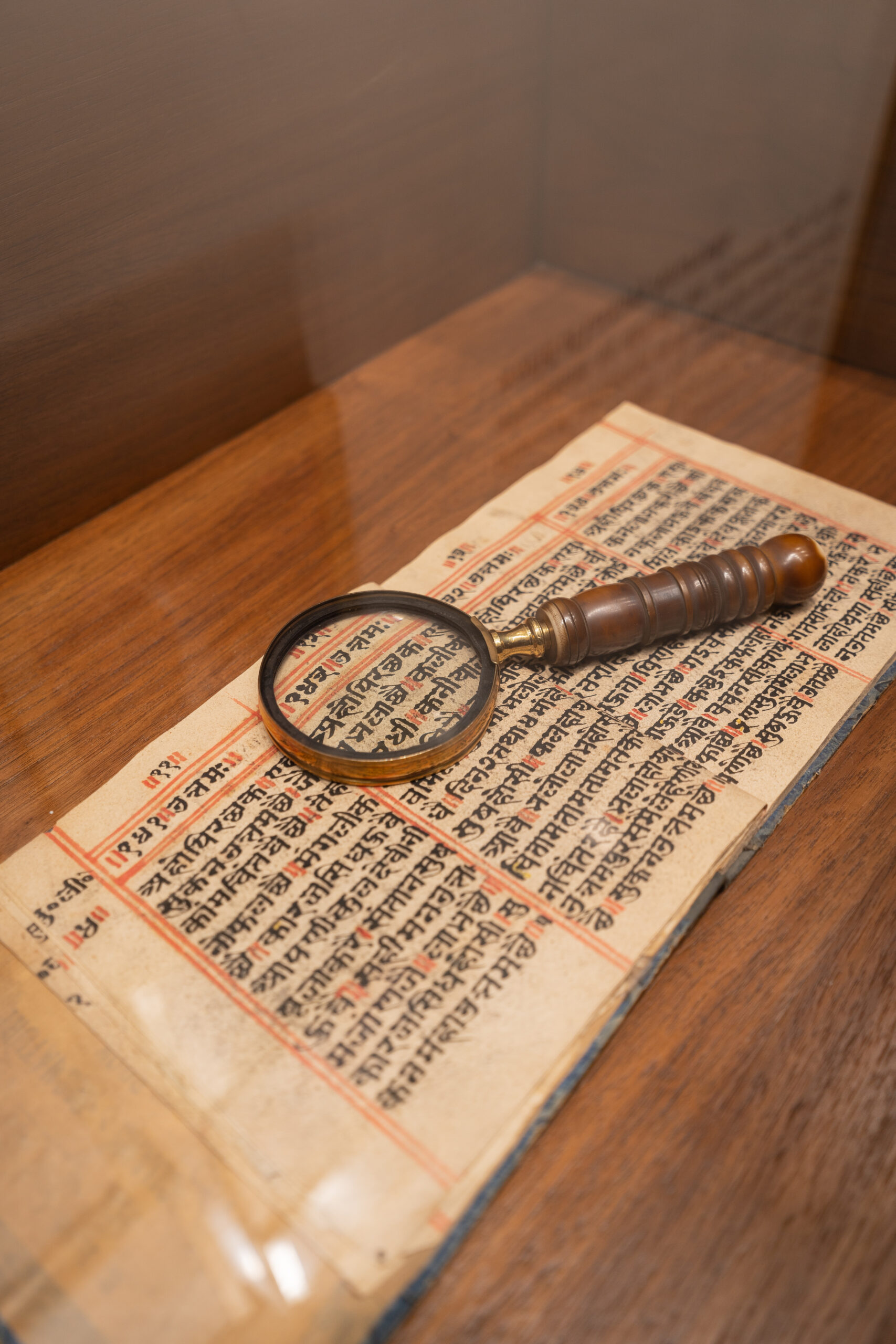 An antique Sanskrit manuscript displayed under glass with a brass magnifying glass resting on it.